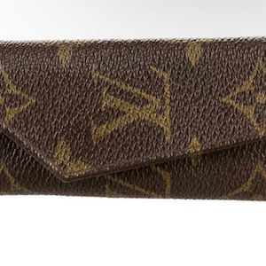 May include: Brown leather key holder with a flap closure. The key holder features a repeating pattern of gold-colored interlocking letters and floral designs. The key holder is rectangular and has a textured surface.
