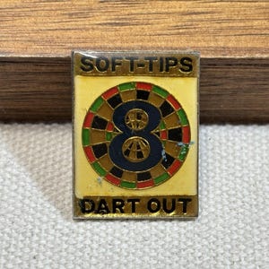 May include: A rectangular pin with the words "SOFT-TIPS" and "DART OUT" in gold lettering. The pin features a dartboard design with a blue "8" in the center, surrounded by red, green, and brown segments on a yellow background.