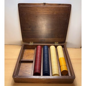 May include: A rectangular wooden box with a hinged lid containing four vertical compartments. Three compartments are filled with stacks of red, blue, and cream-colored circular gaming chips, while the fourth contains a stack of yellow chips.