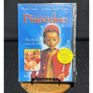 May include: The Adventures of Pinocchio DVD case featuring Martin Landau and Jonathan Taylor Thomas. The cover shows a wooden puppet character in a red jacket, a small inset photo of two people, and a yellow circular sticker highlighting extra features.