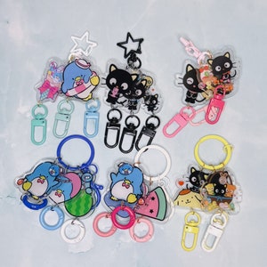May include: A set of six colorful keychains featuring cartoon characters. The keychains are made of clear acrylic and feature different designs, including penguins, cats, and a watermelon. The keychains are attached to metal rings and have different colored charms.