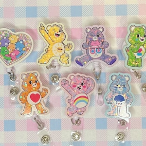 May include: Set of 7 Care Bear themed retractable badge holders. Each holder features a different Care Bear character with glitter accents. The characters include: Funshine Bear, Cheer Bear, Grumpy Bear, Good Luck Bear, Love-A-Lot Bear, Tenderheart Bear, and Wish Bear. The holders are attached to white retractable cords.