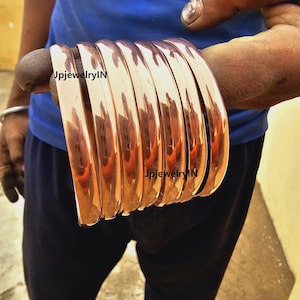 Peut inclure: Une pile de huit bracelets en cuivre, chacun avec une finition lisse et brillante. Les bracelets sont empilés les uns sur les autres, le bracelet du haut étant légèrement décalé par rapport aux autres. Les bracelets sont d'une couleur brun rougeâtre chaude. Le texte "JpjewelryIN" est visible sur deux des bracelets.