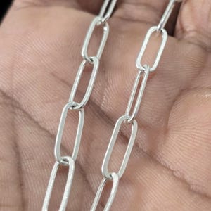 May include: Close-up of two silver-colored chain strands. Each strand is composed of elongated, oval-shaped links. The chains are displayed against a neutral background, highlighting the metallic sheen and the detailed construction of the jewelry.