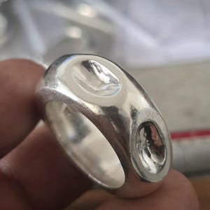 Pode incluir: Um anel de prata com um design oval apresentando duas aberturas arredondadas e rebaixadas. O anel tem uma superfície lisa e polida e uma banda espessa. O anel é segurado pelos dedos de uma pessoa.