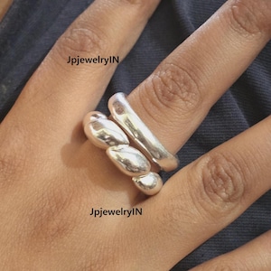 Puede incluir: Dos anillos de plata en un dedo. El anillo superior es una banda simple, mientras que el anillo inferior es un grupo de tres formas redondeadas.