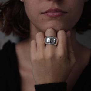 Può includere: Un anello d'argento con una fascia larga e arrotondata. L'anello è liscio e lucido.