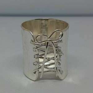 Op de afbeelding: Een zilveren ring met een gedetailleerd ontwerp van een korset met een strik aan de bovenkant.