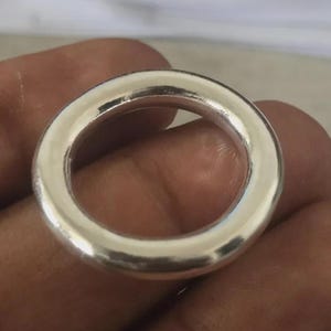 Puede incluir: Un anillo plateado brillante con un diseño liso y redondeado. El anillo es circular y parece estar hecho de metal. El primer plano resalta la superficie pulida y el estilo simple y elegante del anillo. El diámetro del anillo es de aproximadamente 2,5 cm.