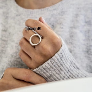 Pode incluir: Um anel de prata com um grande design de círculo aberto. O anel é usado em um dedo.