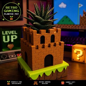 Puede incluir: Una maceta marrón en forma de castillo con una base verde, que alberga una planta suculenta. La maceta tiene ventanas arqueadas y una puerta. El texto en la maceta dice "RETRO GAMING PLANTER POT". Un letrero de "LEVEL UP" está en el fondo.
