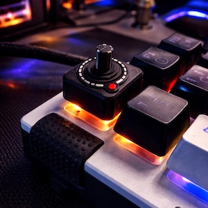 May include: Close-up of a custom mechanical keyboard with a joystick and black keycaps. The keyboard has a silver frame and orange backlighting. A small red light is visible on the joystick.