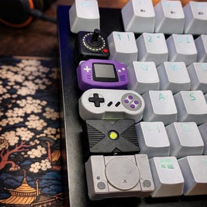 Może przedstawiać: Zbliżenie klawiatury mechanicznej z niestandardowymi nakładkami na klawisze w kształcie kontrolerów do gier retro. Nakładki na klawisze obejmują joystick, fioletową konsolę przenośną, biały kontroler, czarne logo Xbox i szare logo PlayStation.