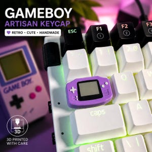 May include: A close-up of a white keyboard with a purple Game Boy artisan keycap. The keycap is shaped like a miniature Game Boy with a screen and buttons. The keyboard also has black keys and the text "GAMEBOY ARTISAN KEYCAP".