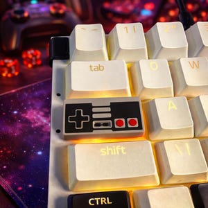 Pode incluir: Close-up de um teclado mecânico com teclas brancas. A tecla da barra de espaço é um design preto e cinza de um controlador de videogame clássico. O teclado tem um tema de jogo retrô.