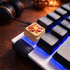 Könnte beinhalten: Eine individuelle Tastenkappe in Form eines Waffelstapels, belegt mit Butter und Speck. Die Kappe befindet sich auf einer weißen und schwarzen mechanischen Tastatur mit blauer LED-Beleuchtung. Kopfhörer sind im Hintergrund zu sehen.