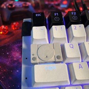 May include: A close-up of a mechanical keyboard with white keycaps. The "caps" key is illuminated with blue light. A keycap shaped like a miniature PlayStation console is centered on the keyboard. Other keys include "ESC", "F1", "F2", "1 !", and "2 ".