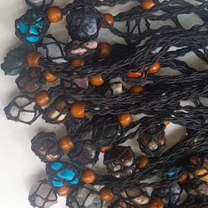 May include: A collection of colourful gemstone pendants, each wrapped in a black macrame net with brown wooden beads. The pendants are displayed on a white surface.