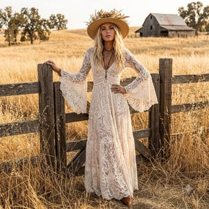 May include: A floor-length cream-colored lace dress featuring long bell sleeves, a deep V-neckline, and a fitted waist. The garment is paired with a wide-brimmed straw hat adorned with dried flowers and a layered necklace with a pendant.