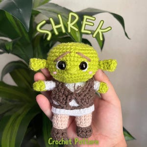 May include: A handmade green crochet Shrek doll with black eyes, small ears, and a brown vest. The doll is wearing a white shirt and tan pants. The word "Shrek" is written in green above the doll, and "Crochet Pattern" is written below.