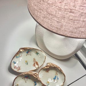 May include: Three decorative seashell dishes with gold-painted edges. Each dish features a whimsical illustration of Peter Rabbit and other characters. The dishes are arranged on a white surface, with a lamp in the background.