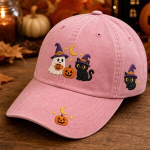May include: A dusty pink baseball cap featuring embroidered Halloween designs. The front displays a ghost in a purple witch hat holding a mug, a jack-o'-lantern, and a black cat in a purple witch hat. A smaller black cat is on the side, with a pumpkin on the brim.