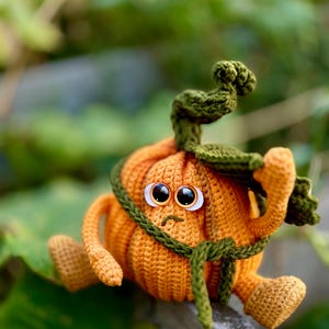 May include: A crocheted pumpkin with a green stem and large eyes, sitting on a gray surface. The pumpkin is wearing a green crocheted belt.