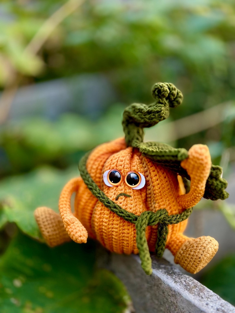 May include: A crocheted pumpkin with a green stem and large eyes, sitting on a gray surface. The pumpkin is wearing a green crocheted belt.