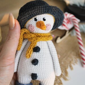May include: A white crocheted snowman with a black hat, yellow scarf, and black buttons. The snowman is wearing blue crocheted boots with white pom-poms.