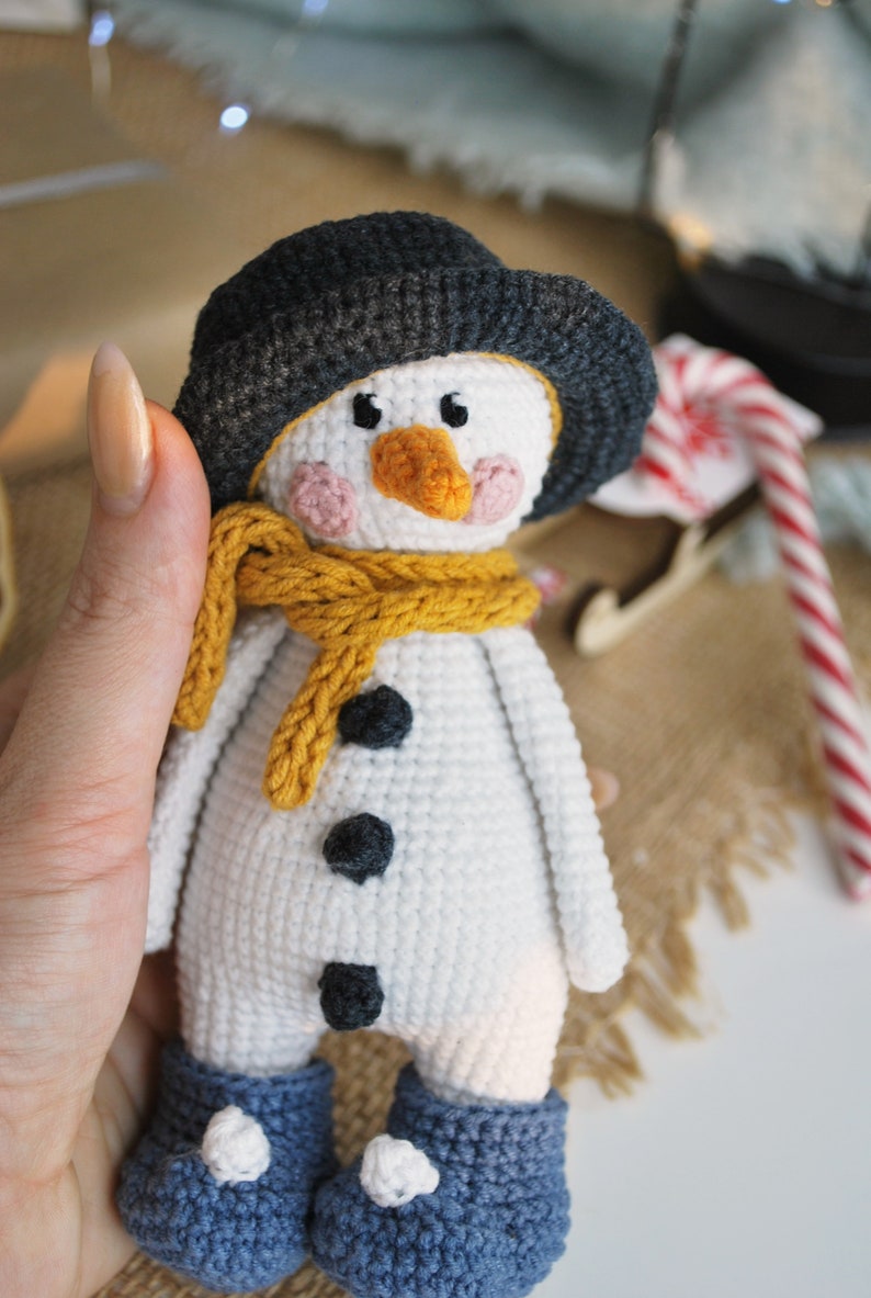 May include: A white crocheted snowman with a black hat, yellow scarf, and black buttons. The snowman is wearing blue crocheted boots with white pom-poms.