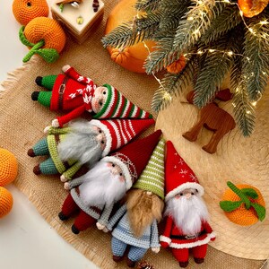 May include: Five crocheted gnomes with different colored hats and outfits. The gnomes are arranged in a semi-circle on a brown burlap surface. The gnomes are wearing red, green, blue, and white hats and outfits.