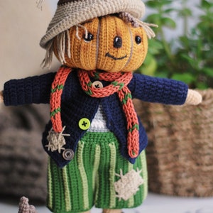 May include: A crocheted pumpkin scarecrow doll wearing a blue jacket, green striped trousers, and a red and green scarf. The doll has a smiling face and is standing next to a small crocheted pumpkin.