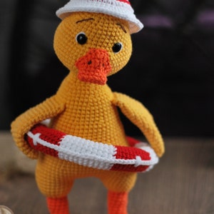 May include: A yellow crocheted duck wearing a white and red crocheted hat and holding a red and white crocheted life preserver.