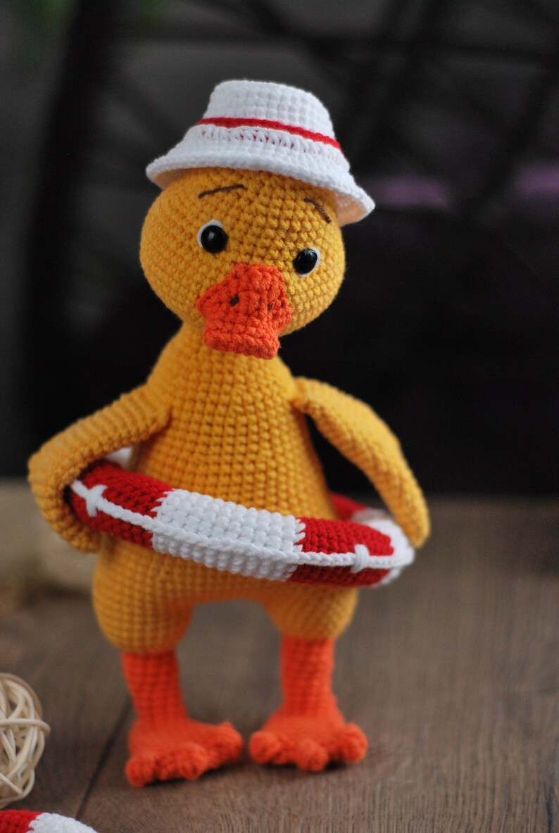 May include: A yellow crocheted duck wearing a white and red crocheted hat and holding a red and white crocheted life preserver.