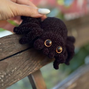 Peut inclure: Une araign&eacute;e crochet&eacute;e marron fonc&eacute; avec de grands yeux brun dor&eacute;. Le jouet en peluche a huit pattes et un corps textur&eacute;. L'araign&eacute;e est pos&eacute;e sur une surface en bois.