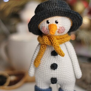 May include: A crocheted snowman toy with a black hat, yellow scarf, and blue boots. The snowman has a white body, black buttons, and a carrot nose.