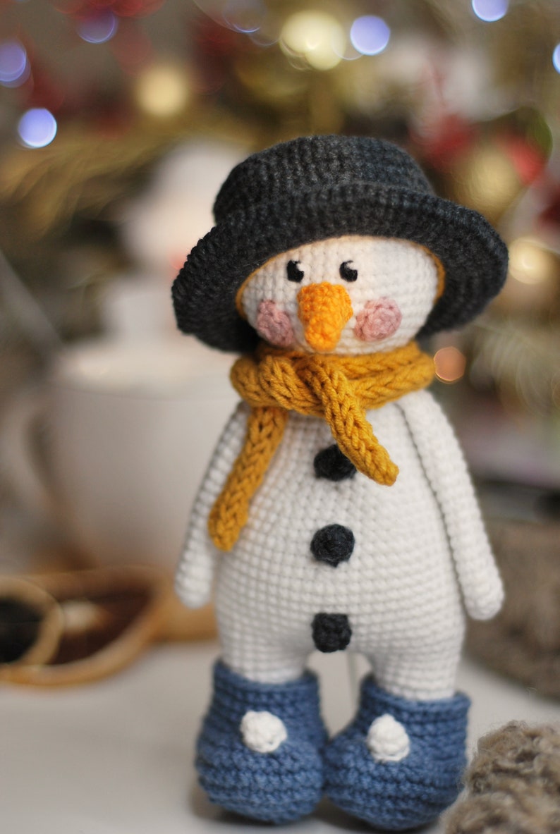 May include: A crocheted snowman toy with a black hat, yellow scarf, and blue boots. The snowman has a white body, black buttons, and a carrot nose.