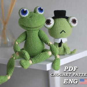Puede incluir: Patrón de crochet para dos juguetes amigurumi de rana verde. La rana más grande tiene ojos azules y articulaciones de madera. La rana más pequeña lleva un sombrero de copa negro y un cuello blanco. El patrón está en inglés e incluye una bandera estadounidense.