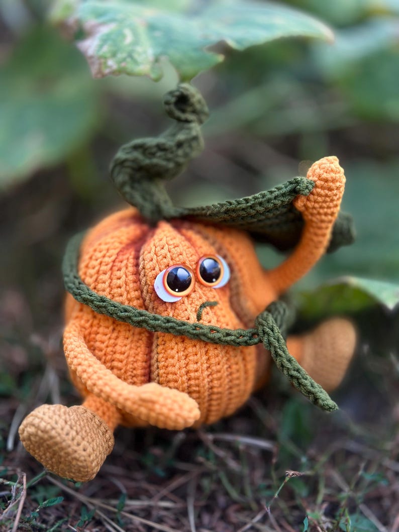 May include: A crocheted pumpkin with a green stem and leaves, wearing a green scarf. The pumpkin has a sad expression with large, round eyes.