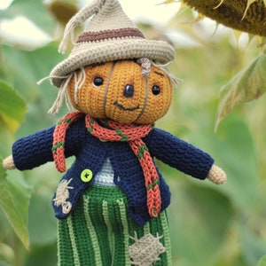 May include: A crocheted pumpkin scarecrow doll wearing a blue jacket, orange scarf, green striped trousers, and a brown hat. The doll is holding a sunflower.