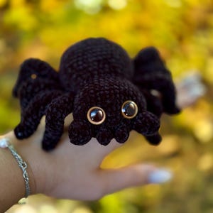 Peut inclure: Une araign&eacute;e noire au crochet avec de grands yeux bruns ronds. L'araign&eacute;e a huit pattes et un corps arrondi. Le jouet en peluche est tenu dans une main sur un fond flou jaune et vert.