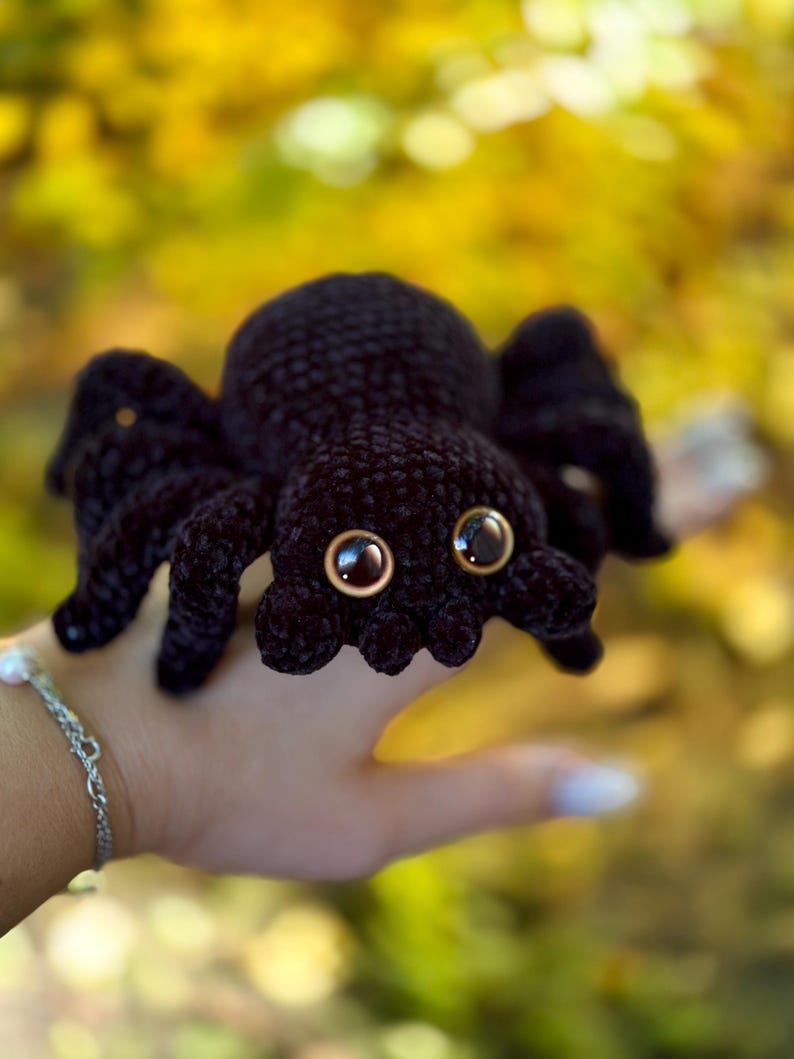 Peut inclure: Une araign&eacute;e noire au crochet avec de grands yeux bruns ronds. L'araign&eacute;e a huit pattes et un corps arrondi. Le jouet en peluche est tenu dans une main sur un fond flou jaune et vert.