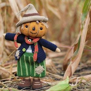 May include: A crocheted pumpkin scarecrow doll wearing a blue sweater, orange scarf, and green striped trousers. The doll is standing in a field of dried corn stalks.