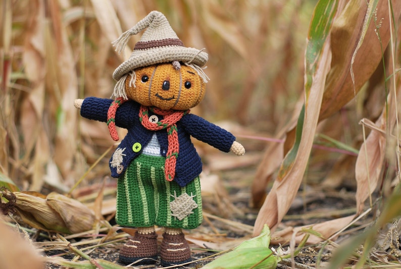 May include: A crocheted pumpkin scarecrow doll wearing a blue sweater, orange scarf, and green striped trousers. The doll is standing in a field of dried corn stalks.