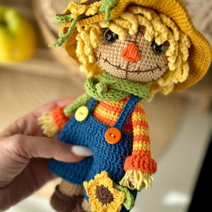 May include: A handmade crochet scarecrow doll with a yellow hat, straw-like hair, and a green scarf. The doll wears blue dungarees with orange and yellow striped sleeves, and holds a sunflower. The doll has a stitched smile and an orange nose.