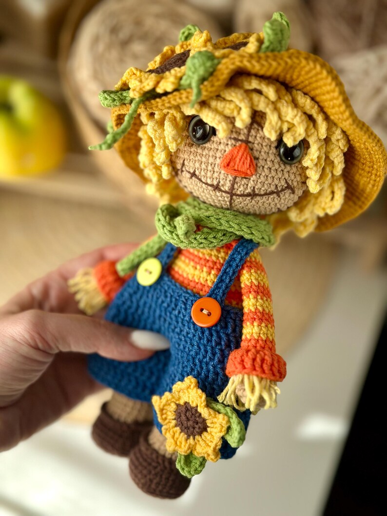 May include: A handmade crochet scarecrow doll with a yellow hat, straw-like hair, and a green scarf. The doll wears blue dungarees with orange and yellow striped sleeves, and holds a sunflower. The doll has a stitched smile and an orange nose.