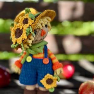 May include: A handmade crochet scarecrow doll wearing a yellow sunflower hat, green scarf, and blue dungarees. The doll has an orange nose, brown shoes, and is holding a sunflower. The doll is approximately 15 cm tall.