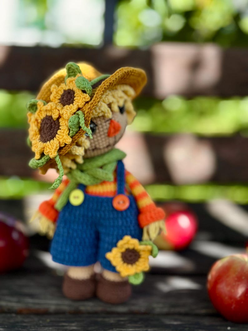 May include: A handmade crochet scarecrow doll wearing a yellow sunflower hat, green scarf, and blue dungarees. The doll has an orange nose, brown shoes, and is holding a sunflower. The doll is approximately 15 cm tall.