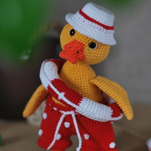 May include: A crocheted yellow duck wearing a white and red hat, red and white polka dot swim trunks, and a red and white life preserver.