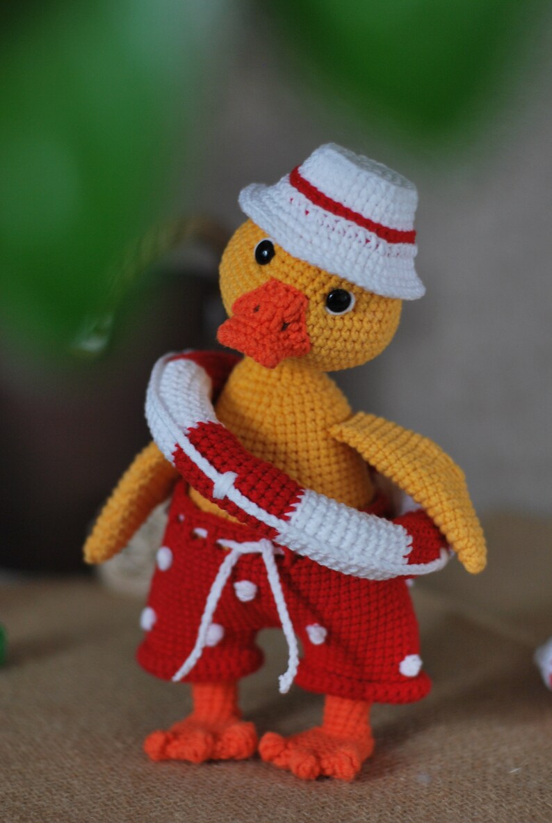May include: A crocheted yellow duck wearing a white and red hat, red and white polka dot swim trunks, and a red and white life preserver.